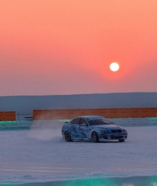 A car is drifting on the ice with the sun setting behind it, creating a stunning scene.