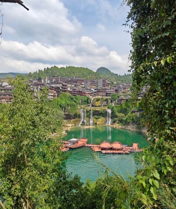 From a distance, the grand waterfall of Furong Ancient Town is framed by emerald-green lake water, with a wooden viewing platform above it.